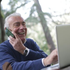 Happy senior man talking on phone and using laptop outdoors.